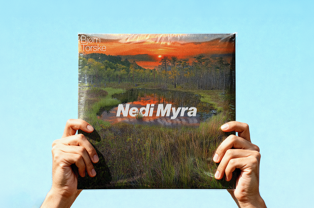Hands holding a vinyl record cover showing a sunset over a marsh with text Nedi Myra and Bjørn Torske.