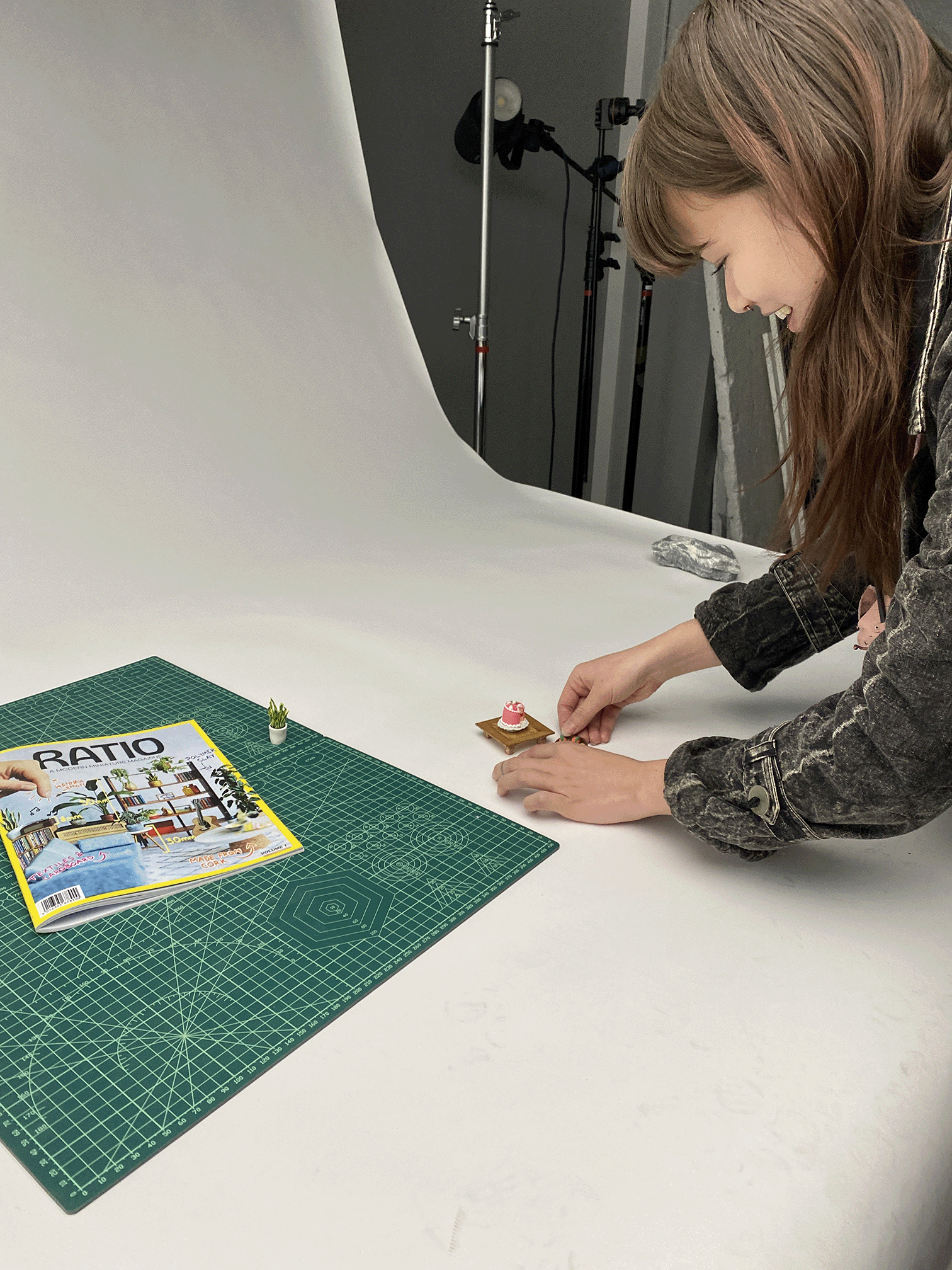 Woman smiling while arranging miniature cake on wooden stand on white tabletop with magazine nearby.