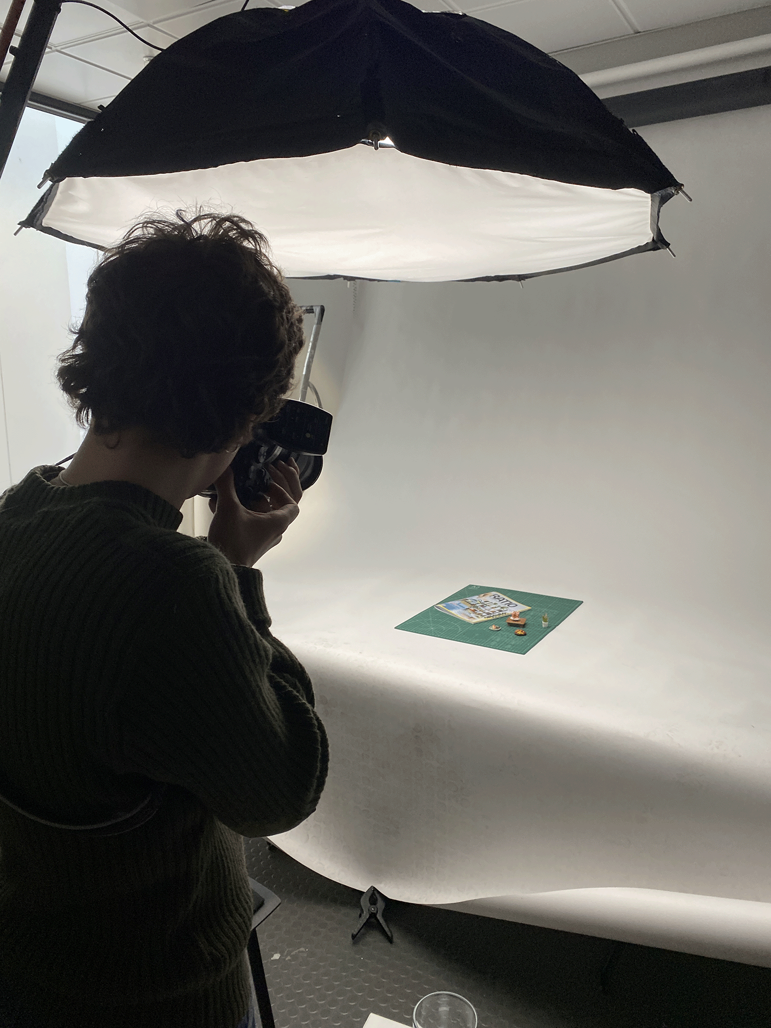 Person photographing small objects and magazine on a green mat under studio lighting.