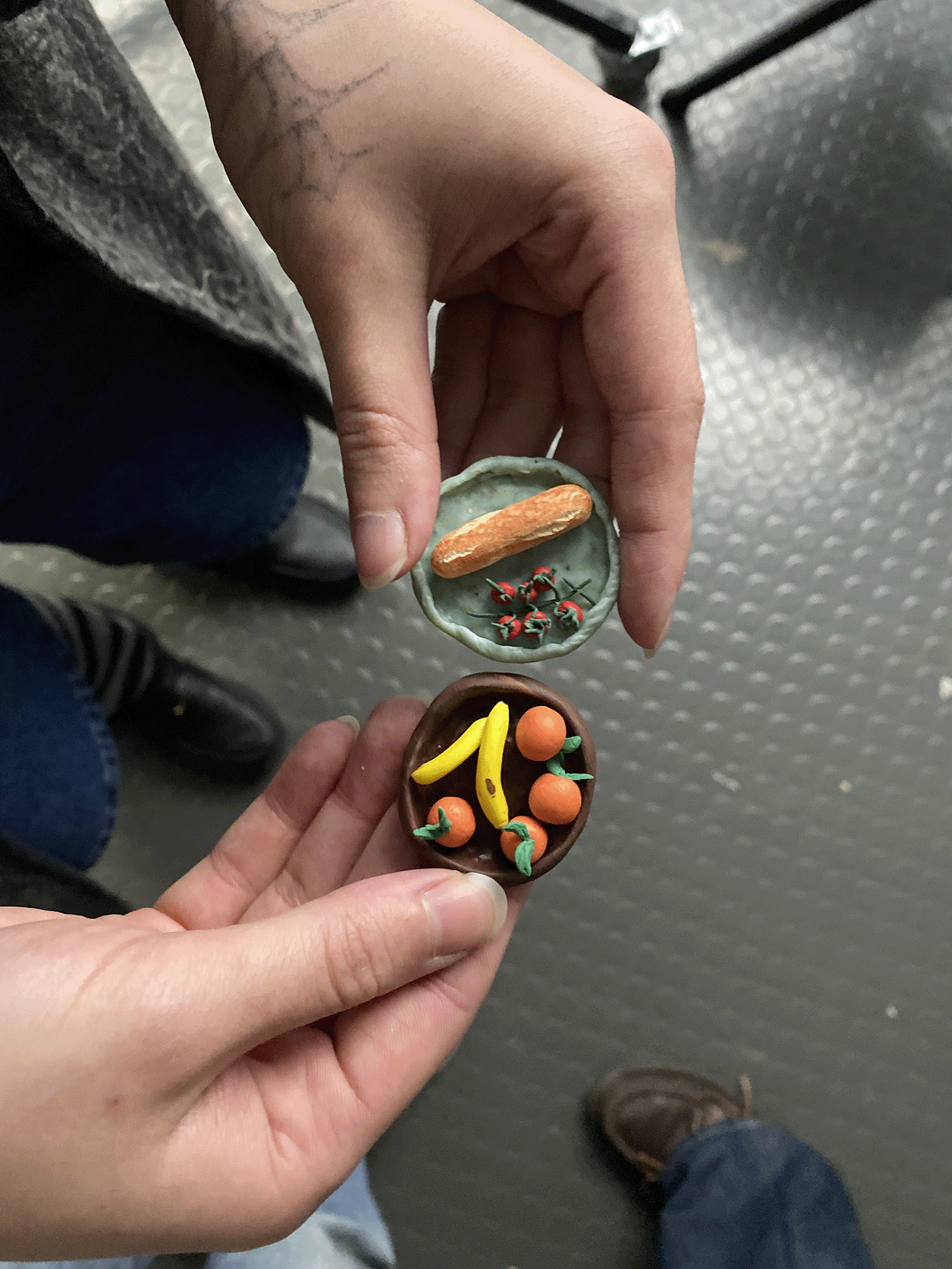 Two hands holding small clay plates with miniature loaf, tomatoes, bananas, and oranges.