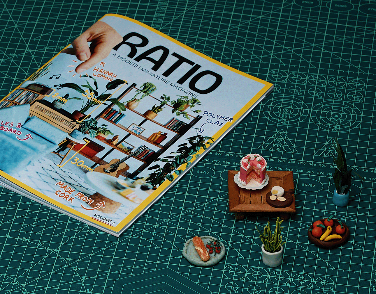 Miniature magazine on green grid cutting mat with tiny plants, cake, fruit, and bread models nearby.