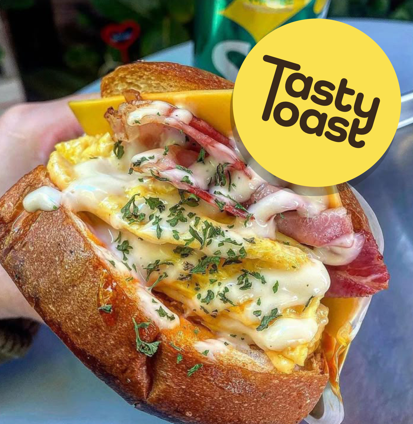Hand holding a toasted sandwich with scrambled eggs, bacon, cheese, and herbs with creamy sauce.