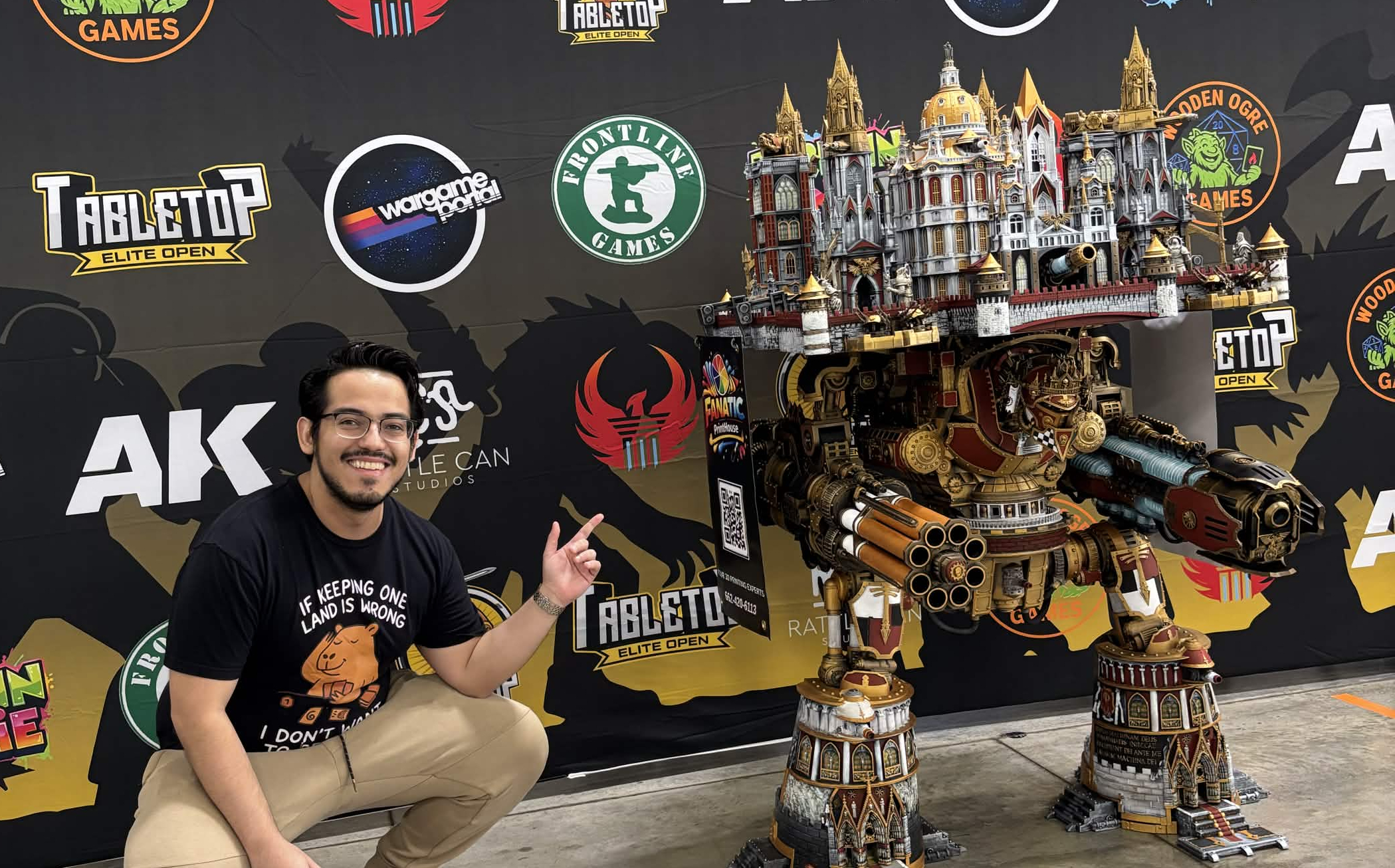 Smiling man wearing glasses, beige pants, and a black t-shirt points at a large, detailed model of a steampunk-style robot with castle-like structures on top, crouching in front of a backdrop with various gaming logos.