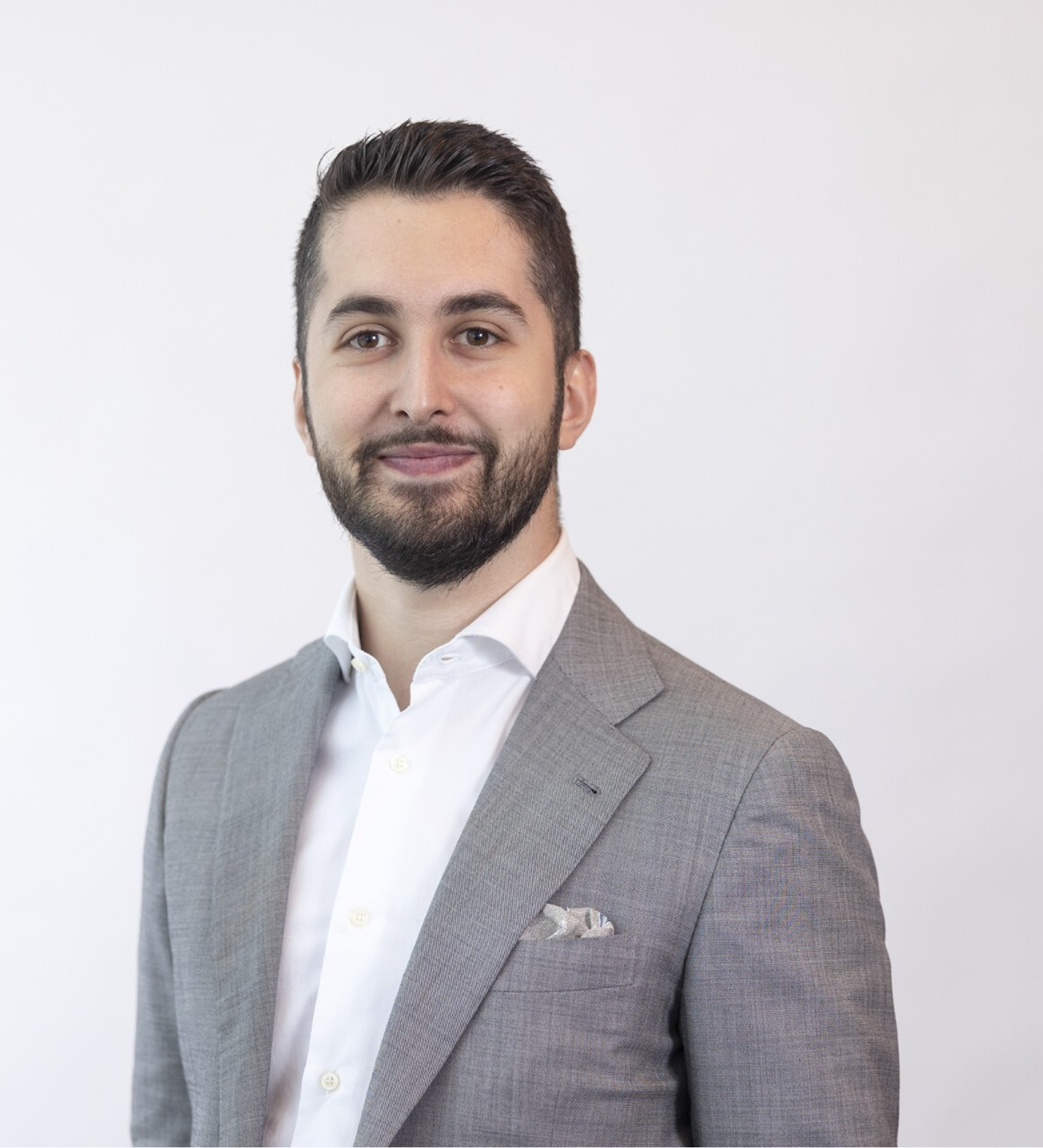 Smiling man with dark hair and beard wearing a gray suit jacket and white shirt against a plain light background.