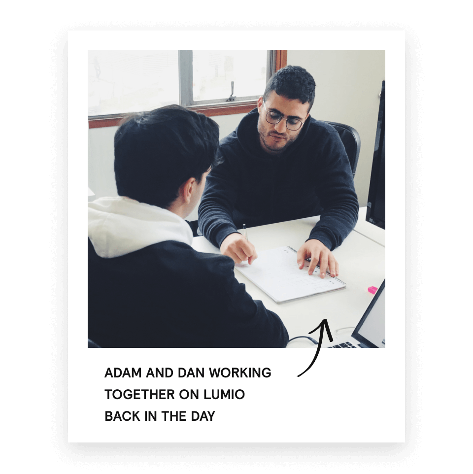 Two men sitting at a desk in an office, one writing in a notebook while the other watches.