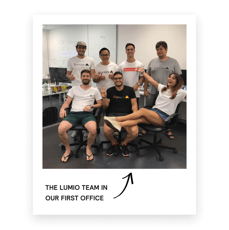 Seven young adults in casual clothes posing and smiling in an office with computers and a whiteboard, some wearing foenix-branded T-shirts.