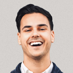 Smiling young man with dark hair wearing a white shirt and dark sweater against a light background.