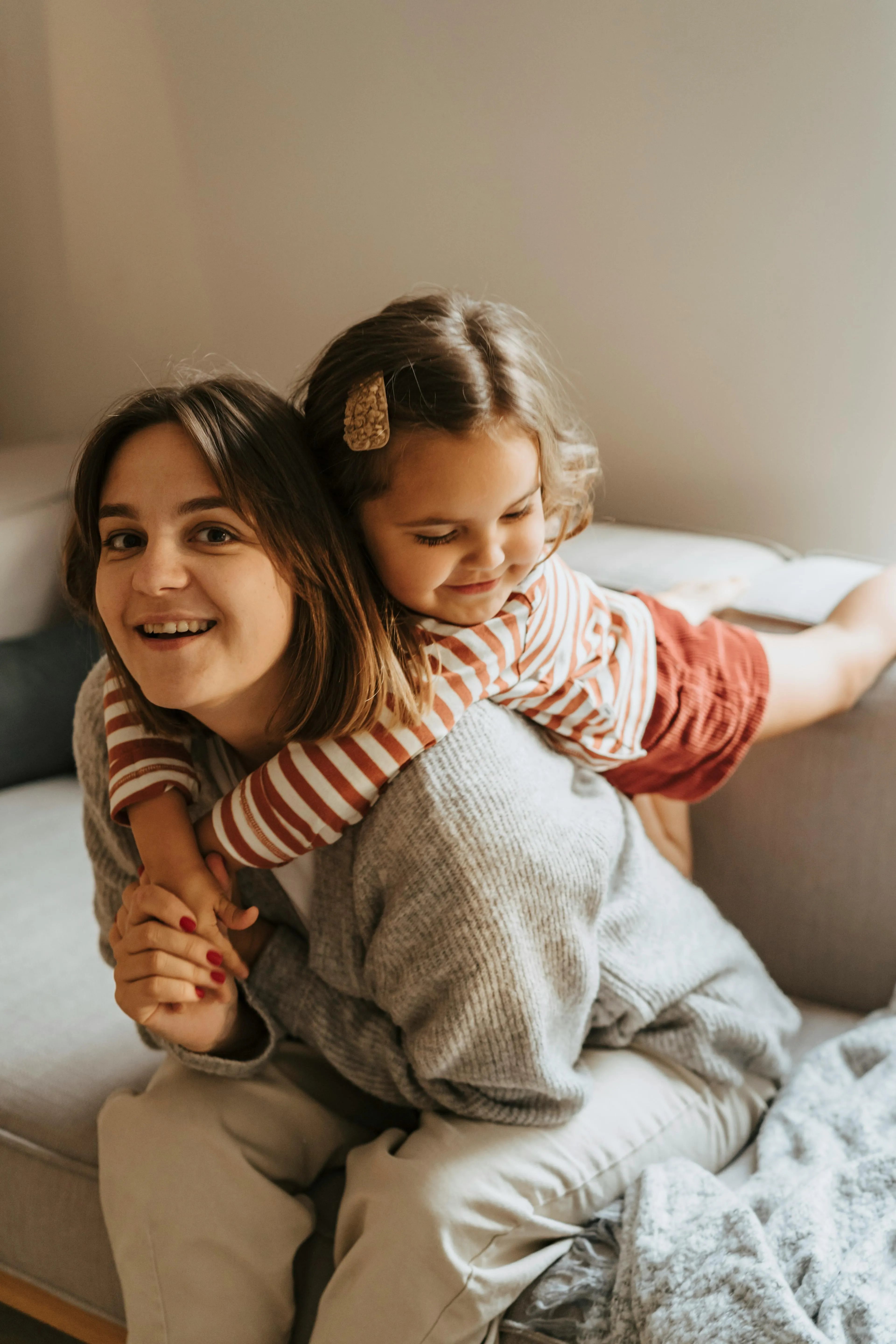 Smiling woman sitting on a couch with a young girl hugging her from behind, both wearing casual clothes.