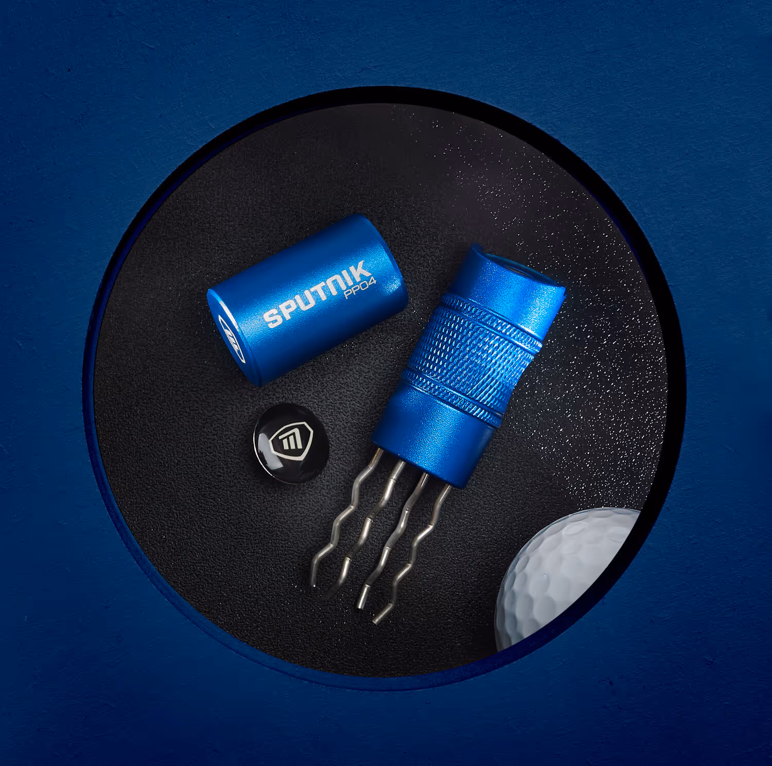 Blue Sputnik pitch repair tool with three metal prongs and a golf ball next to it on a dark surface.