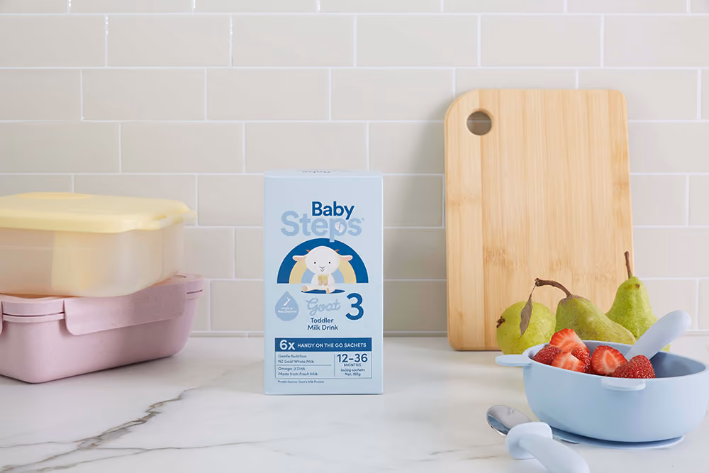Box of Baby Steps Toddler Milk Drink with fruit bowl, pears, cutting board, and storage containers on marble counter.