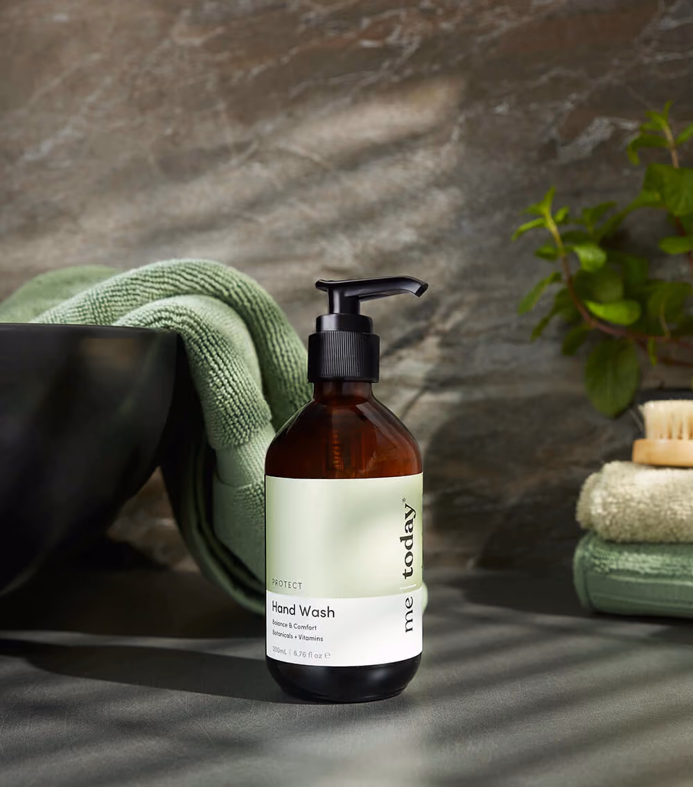 Amber bottle of Me Today hand wash with black pump on table with green towel and plant.