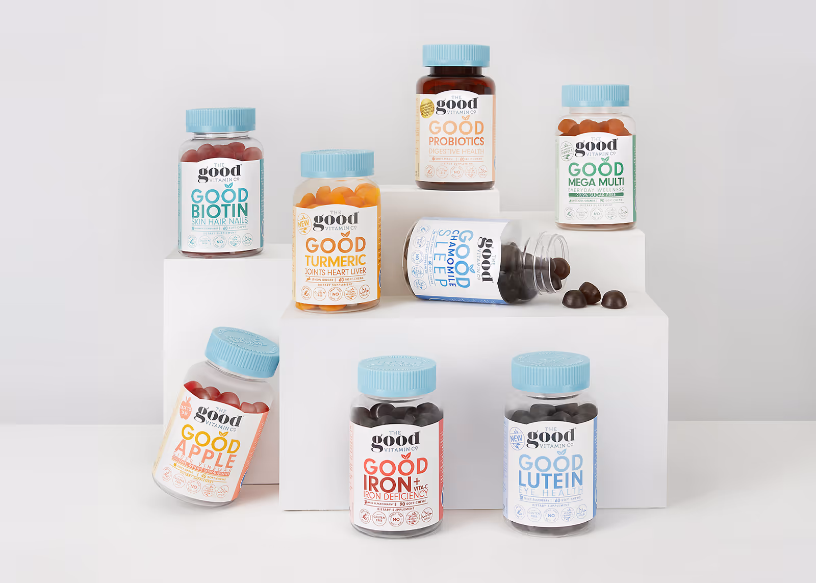 Nine bottles of The Good Vitamin Co supplements arranged on white blocks against a light background.