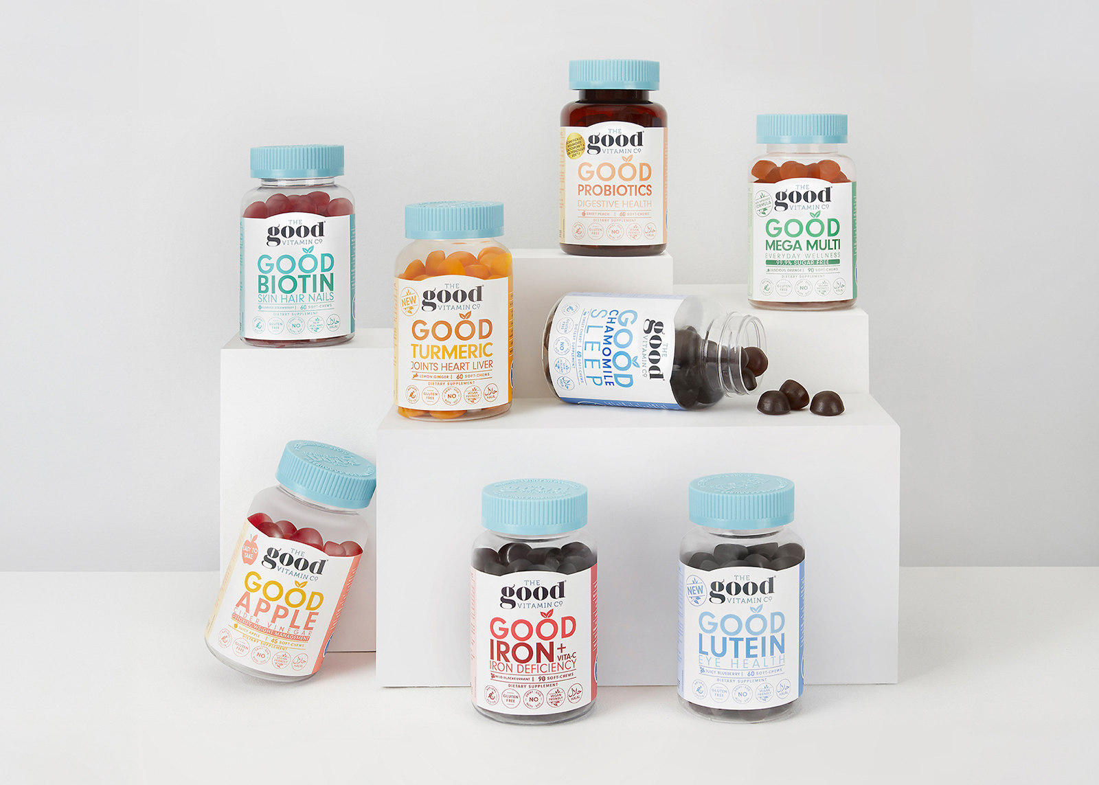 Nine bottles of The Good Vitamin Co supplements arranged on white blocks against a light background.