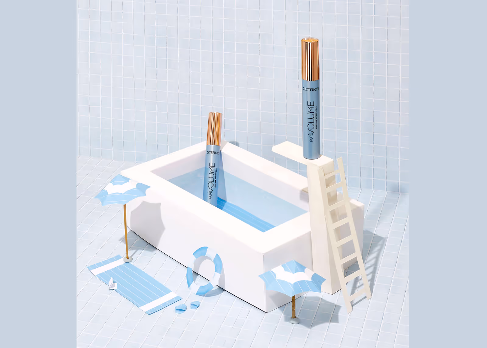 Two mascara tubes styled as swimmers near a mini pool, with diving board, umbrellas, and floaties.