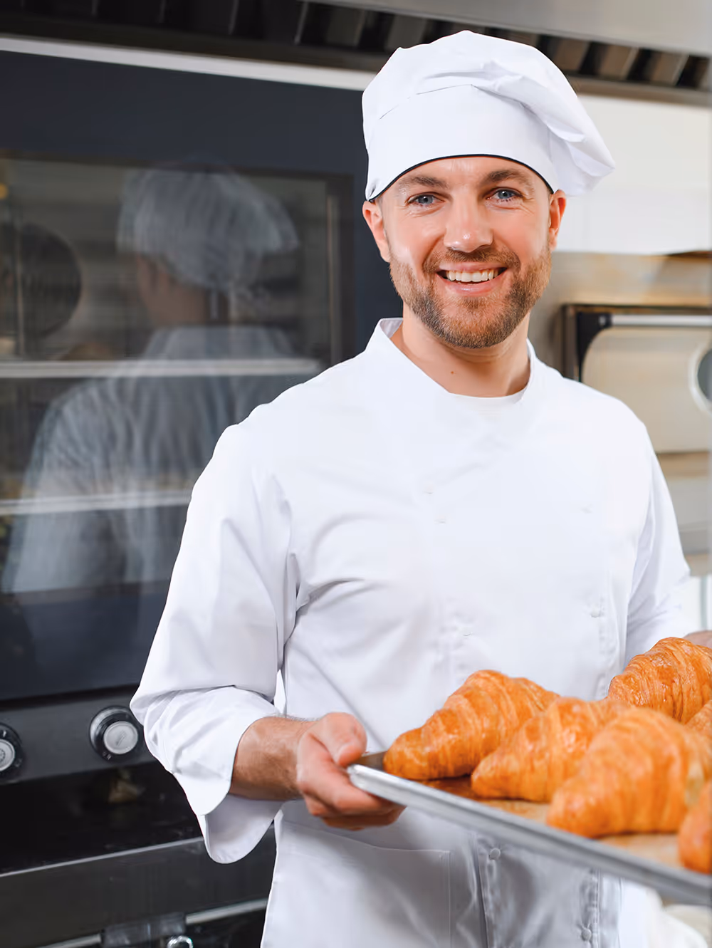 Smiling baker in white uniform holding a tray of fresh croissants in a bakery kitchen.