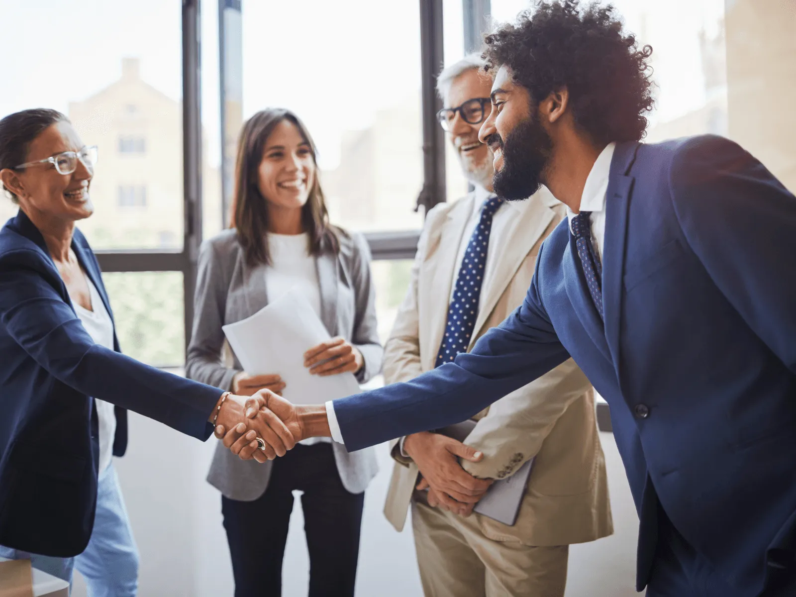 Two business people smiling and shaking hands while two colleagues observe and smile in an office setting.