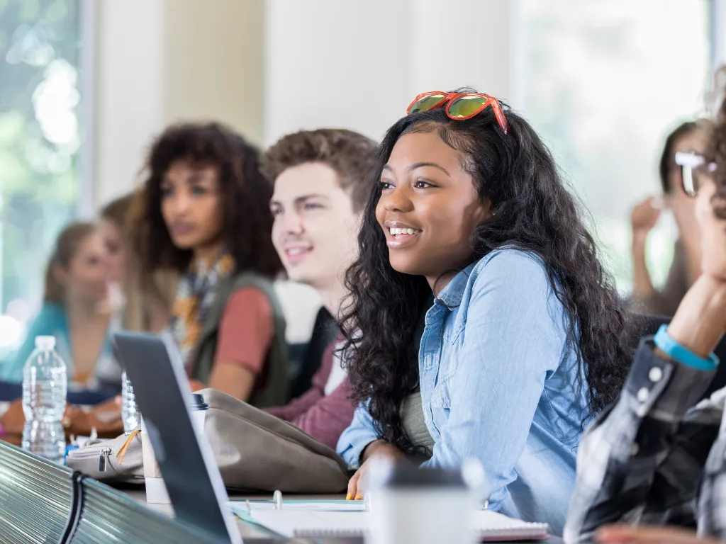 Group of diverse students sitting in a classroom, smiling and engaging with each other, with laptops and notebooks on the desk.