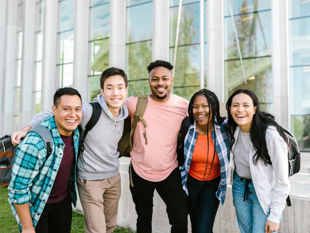 A group of five diverse young adults standing closely together outdoors, smiling and posing for the camera in front of a modern glass building.