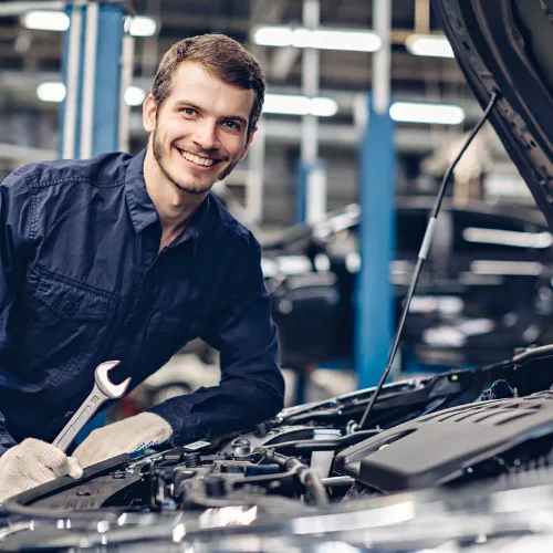 Mechanic smiling while holding a wrench and working on a car engine in a garage.
