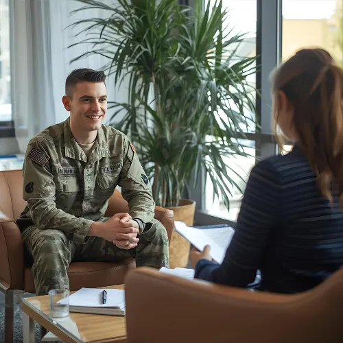 A smiling soldier in military uniform sitting in a chair, talking to a woman who is holding a clipboard in an office setting.