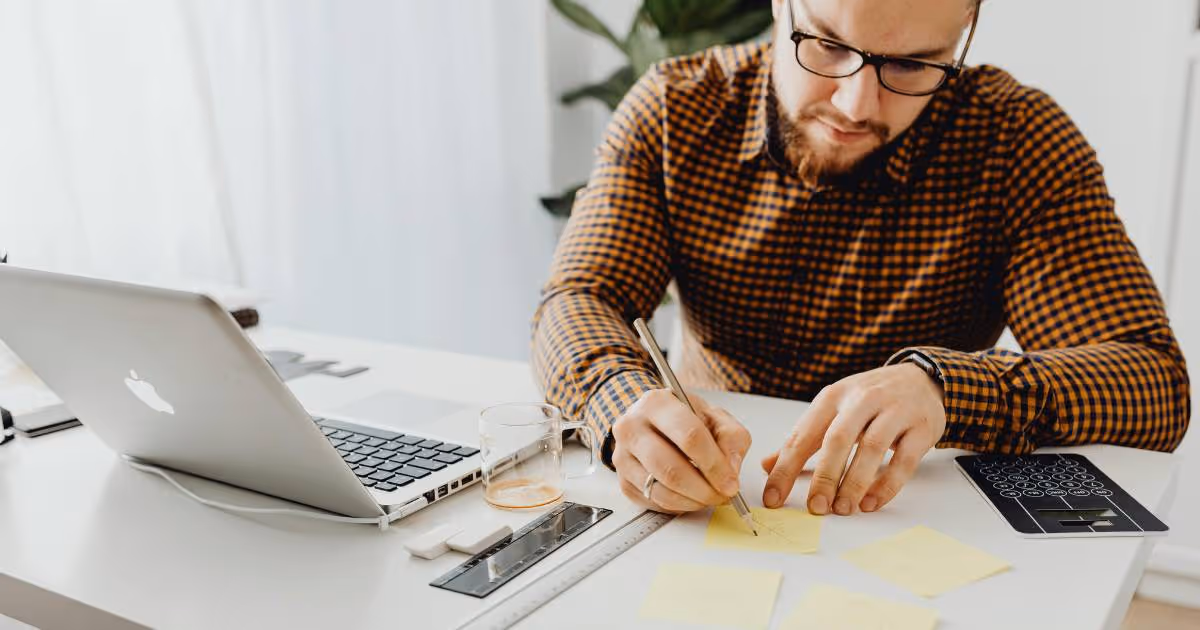 Man at his desk with laptop in front of him while he's writing notes.