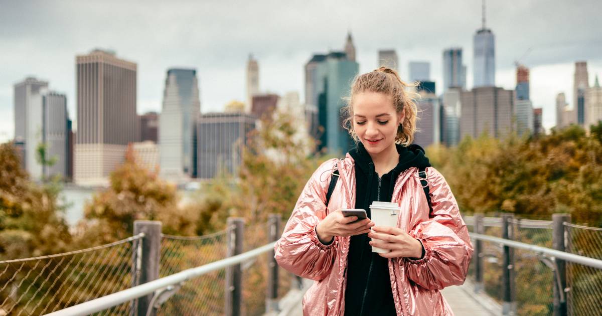 Young woman walking through a city while looking down at her phone, with the city buildings in the background.