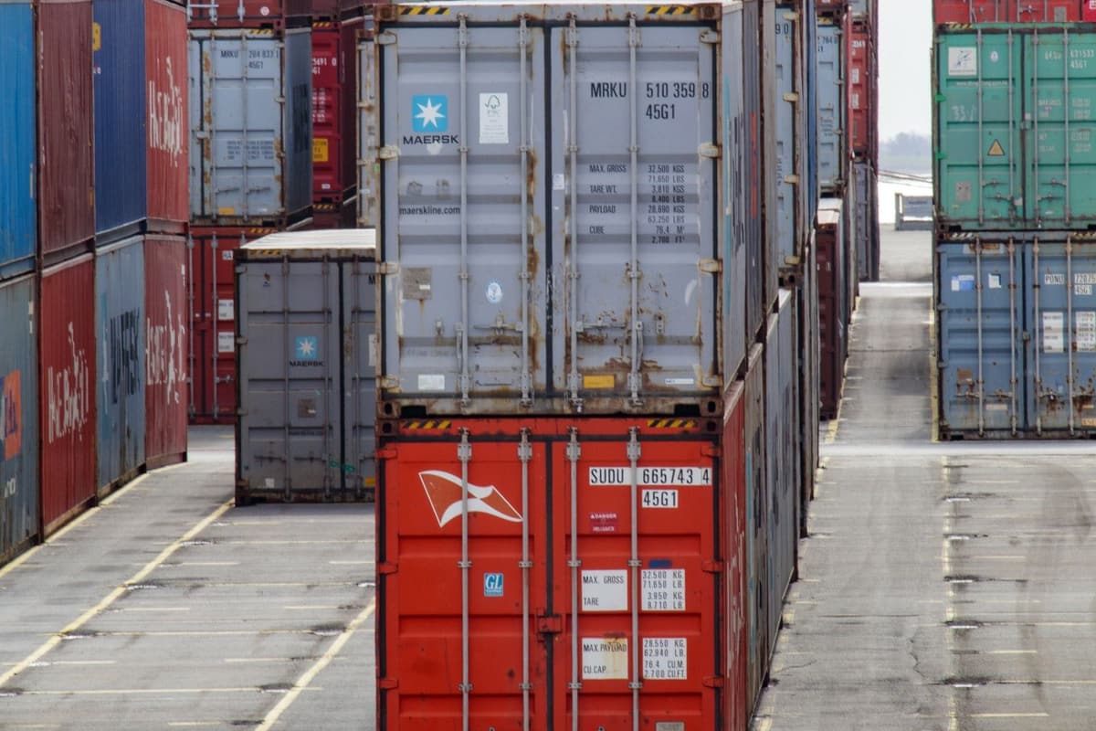 Rows of stacked shipping containers seen head on in a terminal yard, empty marked lanes either side
