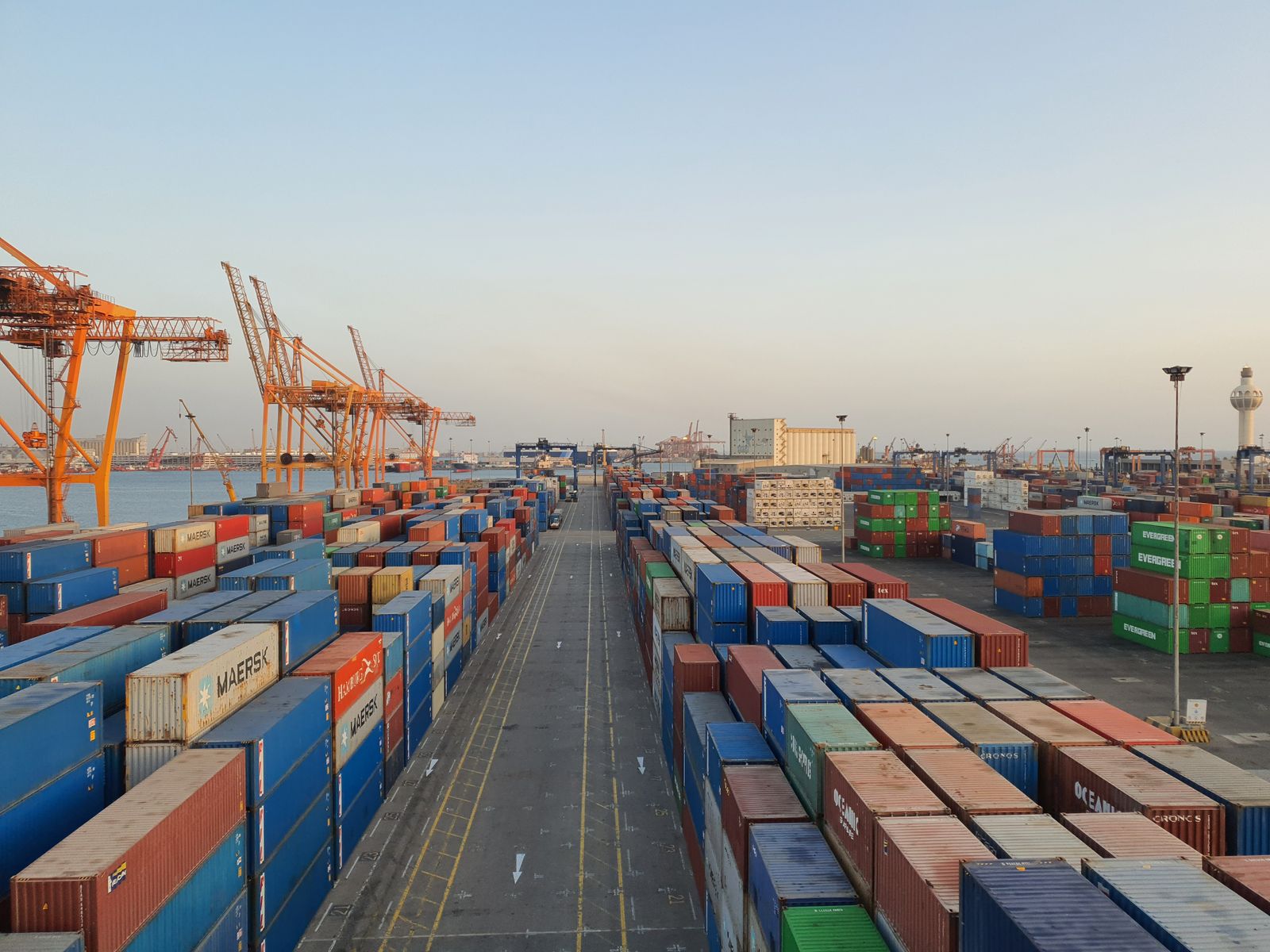 Long rows of stacked containers and quayside cranes at a port terminal at dusk, roadway empty