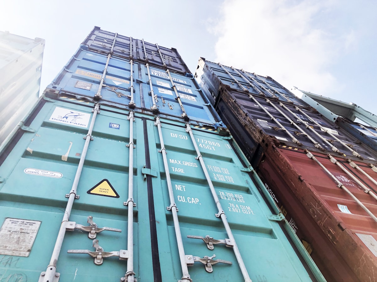 Shipping containers stacked several high against the sky, lashing rods running up the door faces