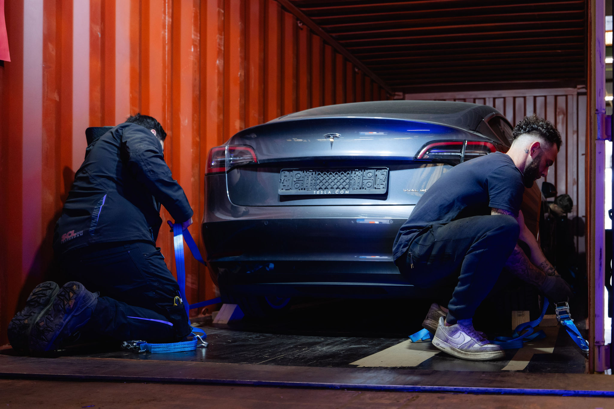Two workers strapping a dark blue electric car down inside a shipping container
