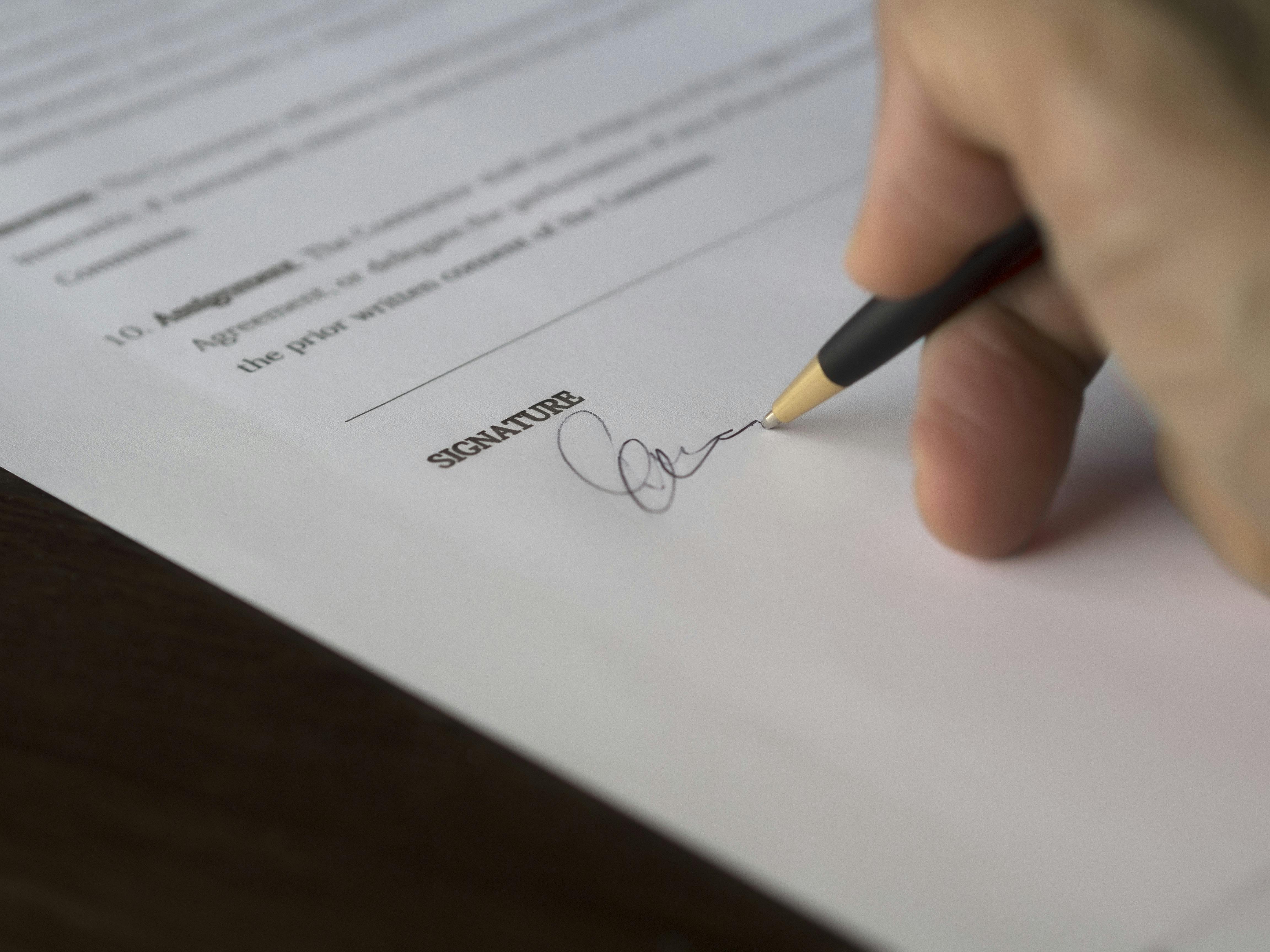 A hand signing a printed contract on the signature line with a pen