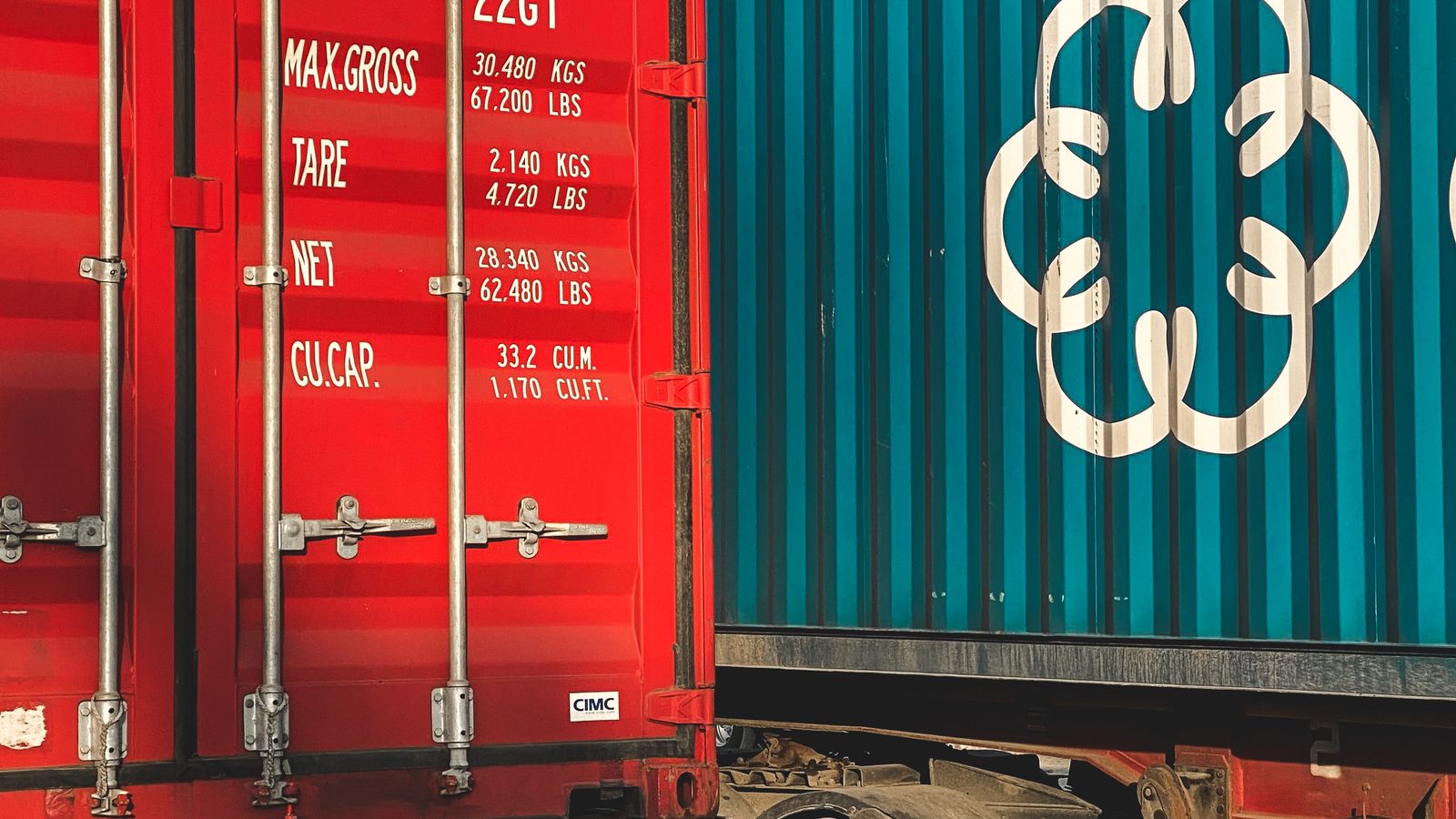 The doors of a red shipping container beside a teal one, weight and capacity markings visible