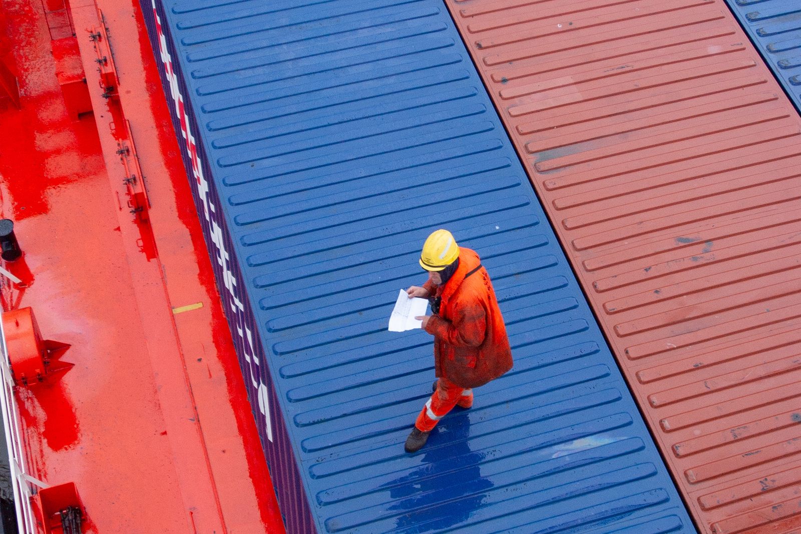 A port worker in a high visibility suit and hard hat checks paperwork while standing on top of stacked shipping containers, viewed from directly above.