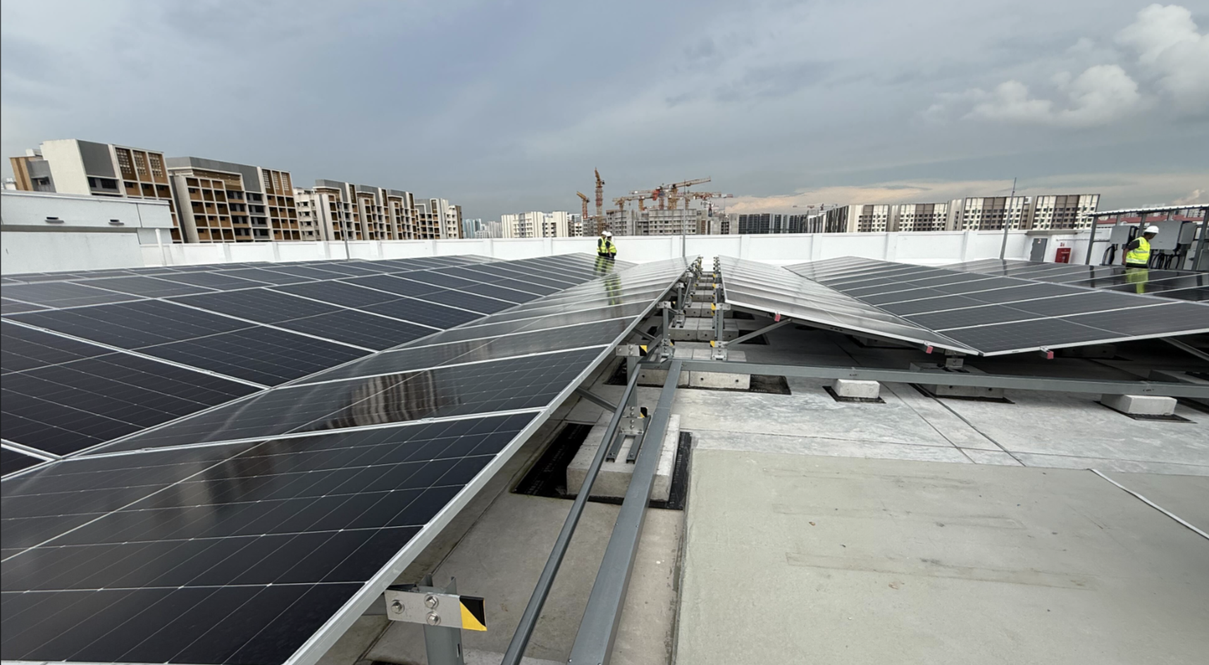 Rows of tilted solar panels on concrete footings across a flat rooftop in Singapore, with installers in safety vests and HDB blocks in the background.