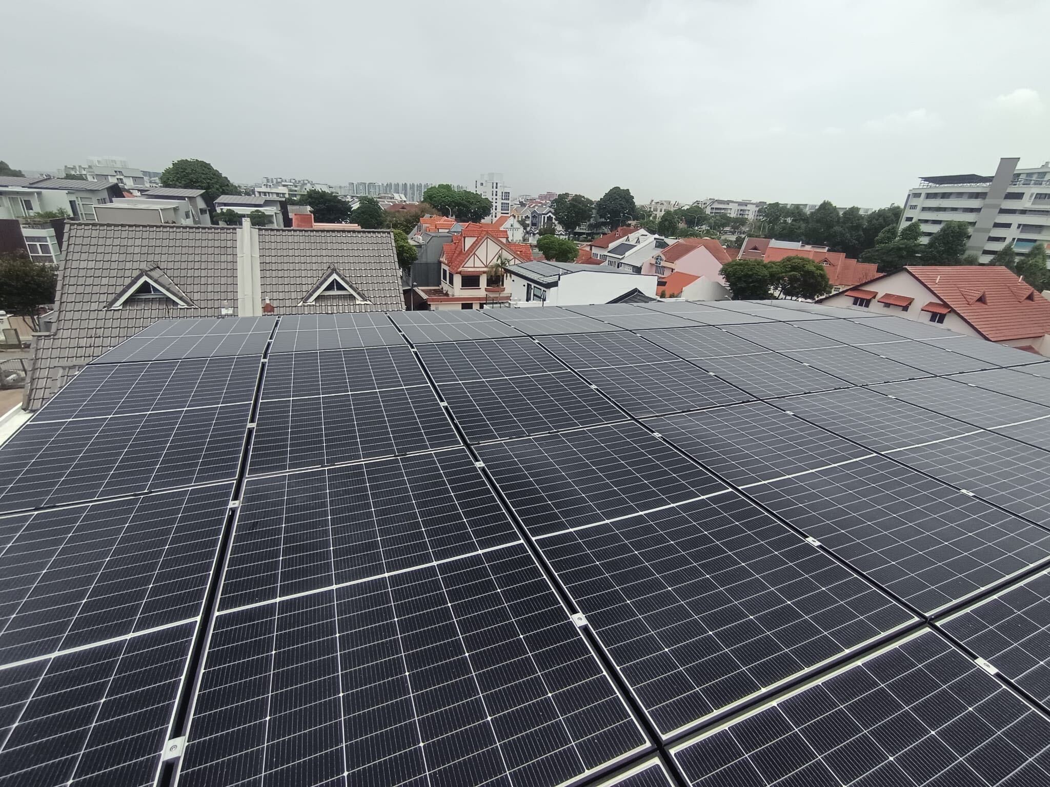 Solar panels on a landed home rooftop in Singapore under a grey, hazy sky, with neighbouring pitched roofs behind