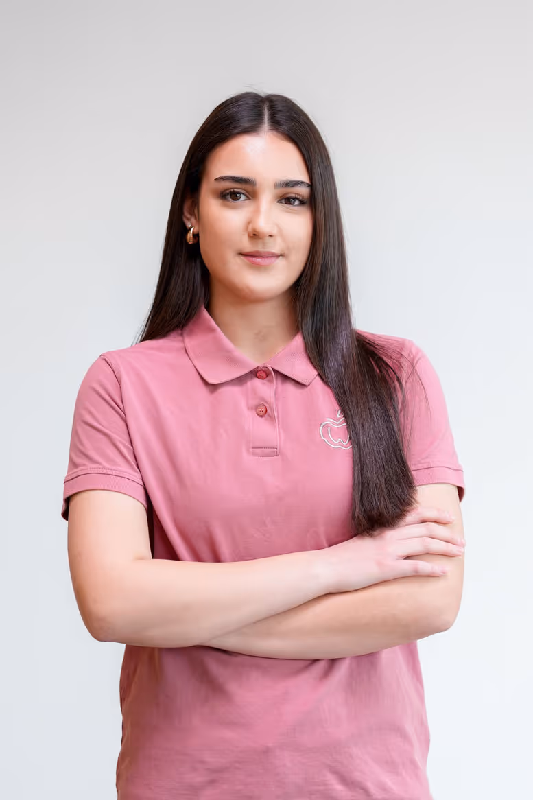 Junge Frau mit langen dunklen Haaren trägt ein rosa Poloshirt und hat die Arme vor der Brust verschränkt.