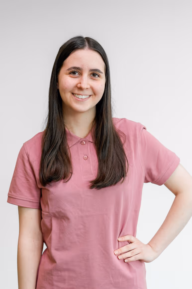 Junge Frau mit langen braunen Haaren trägt ein rosa Polohemd und lächelt, während sie eine Hand in die Hüfte stemmt.