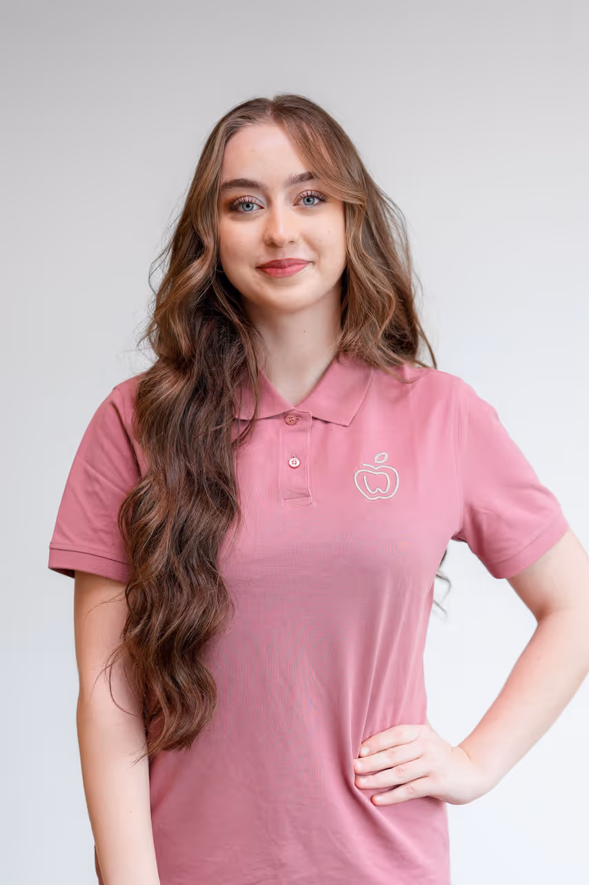 Junge Frau mit langen, welligen braunen Haaren trägt rosa Polohemd mit Apfel-Logo und lächelt freundlich.