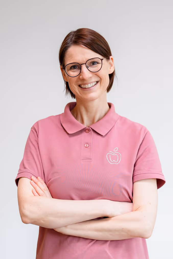 Lächelnde Frau mit kurzen braunen Haaren und Brille, die rosa Polohemd mit Apfel-Logo trägt und die Arme verschränkt.