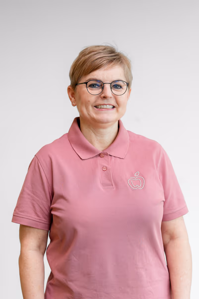 Lächelnde Frau mit kurzen blonden Haaren und Brille trägt ein rosa Polohemd mit einem Apfel-Logo.