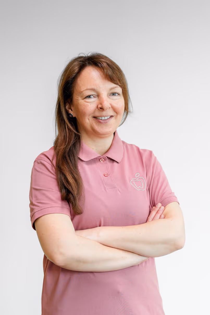 Frau mit braunen Haaren in einem rosa Poloshirt vor weißem Hintergrund, die die Arme verschränkt und lächelt.