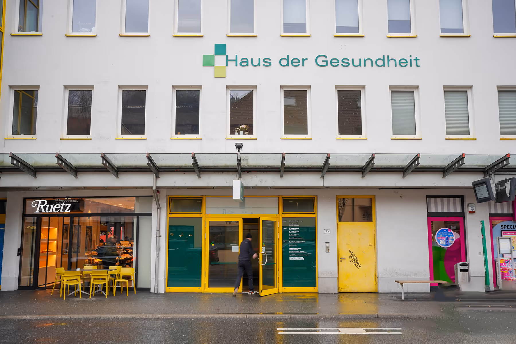 Eingang des Gebäudes Haus der Gesundheit mit gelben Türen und Café mit gelben Stühlen nebenan.