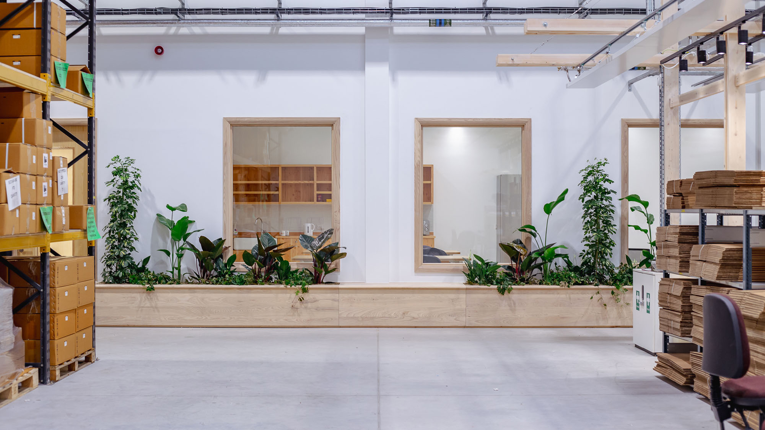A modern warehouse interior for Secure Systems, featuring boxed shelving, long wooden benches with integrated planters, and lush tropical plants throughout the space.