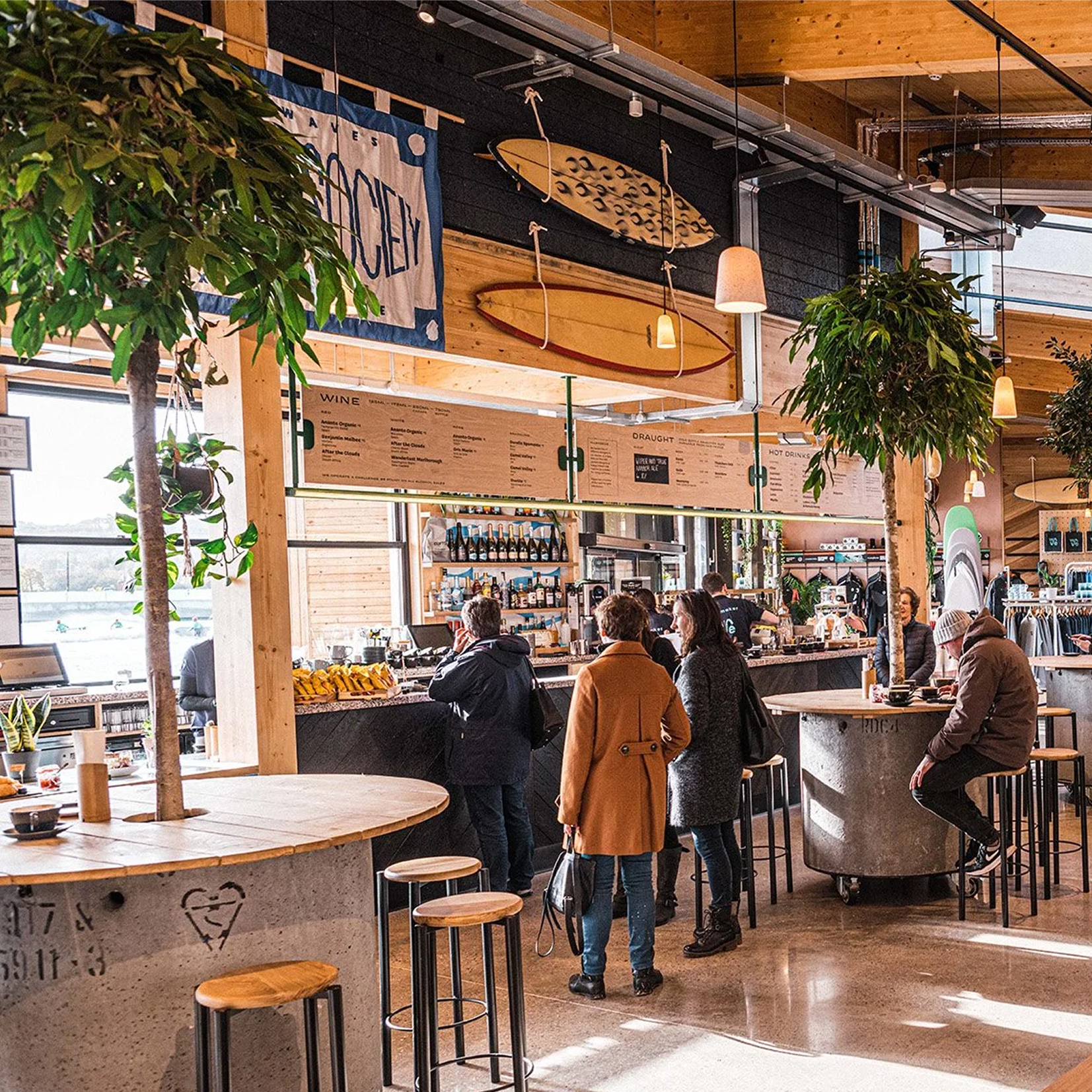 People inside a bright wooden café with surfboards, indoor trees, and a bar counter with stools.
