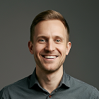 Smiling man with short light brown hair wearing a dark gray collared shirt against a plain gray background.