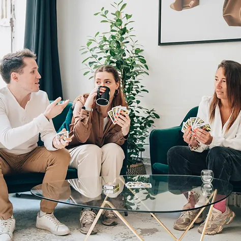 Three people sitting on couches playing cards, with one person drinking from a mug, around a glass coffee table in a living room.
