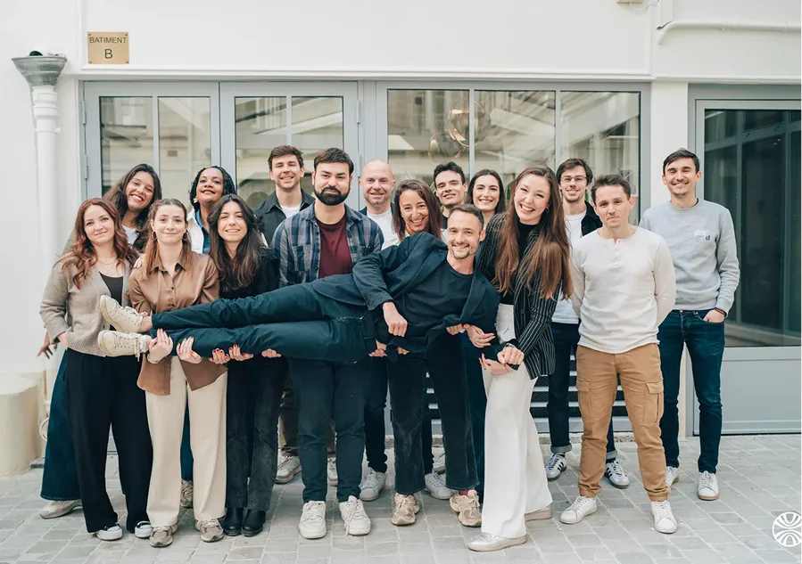 Group of fifteen smiling young adults, with three people holding a man horizontally in front of them, standing outside a building with glass doors.
