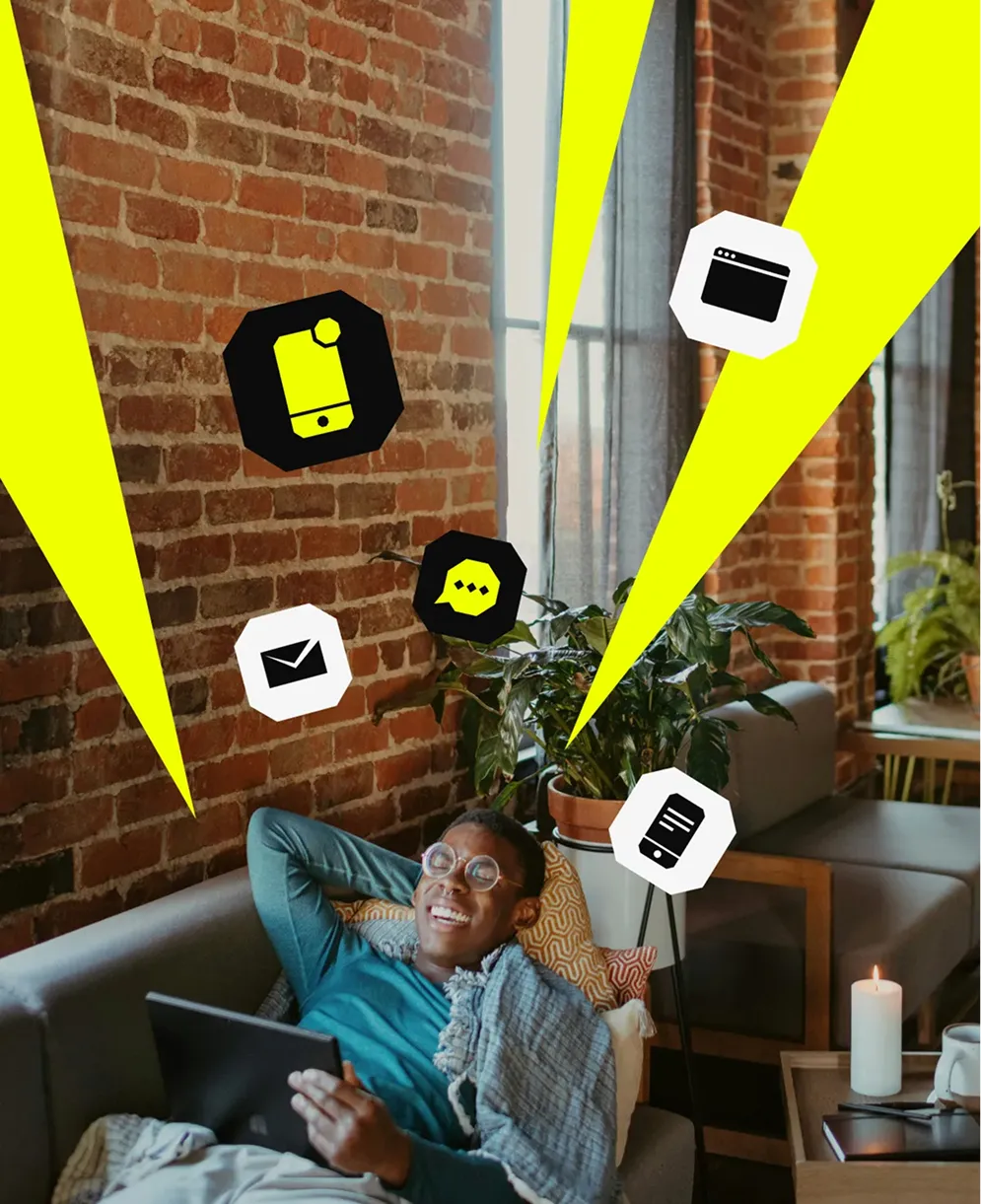 Smiling man lying on couch holding a tablet with digital icons for phone, email, chat, webpage, and document around him in a cozy brick-walled room.