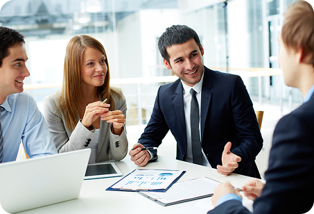 Four business professionals engaged in a discussion around a table with laptops, documents, and a tablet.