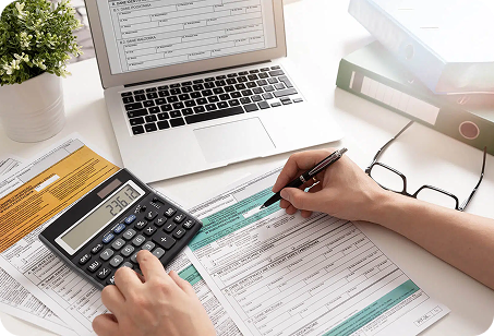 Person filling out forms with a pen while using a calculator and laptop on a desk with glasses and a small plant nearby.